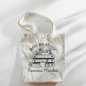 Pu&ograve; includere: Una borsa di tela bianca con testo nero che dice "Meet me at the Farmers Market". Il testo &egrave; circondato da un'illustrazione nera di una cassa di prodotti freschi.