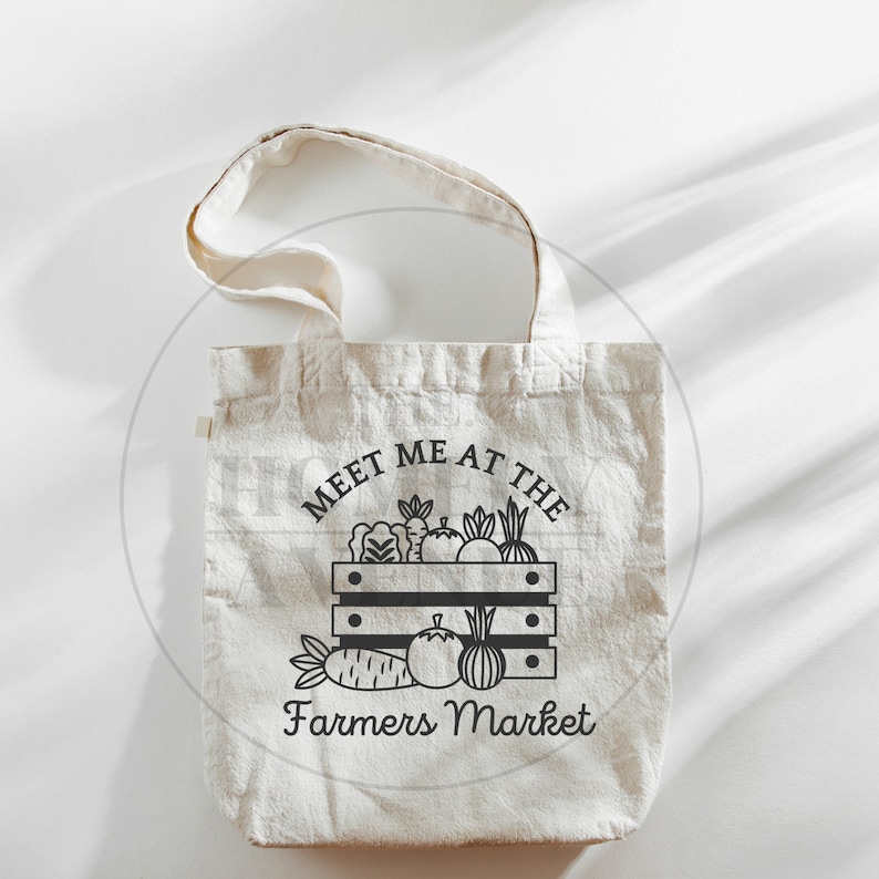 Pu&ograve; includere: Una borsa di tela bianca con testo nero che dice "Meet me at the Farmers Market". Il testo &egrave; circondato da un'illustrazione nera di una cassa di prodotti freschi.