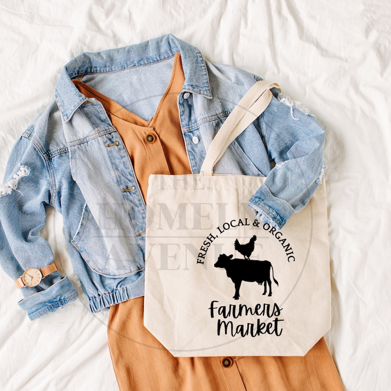 Pu&ograve; includere: Una borsa di tela bianca con testo nero che recita "Fresco, locale & biologico" e "Mercato degli agricoltori". Una silhouette nera di una mucca con una gallina sulla schiena &egrave; al centro della borsa.