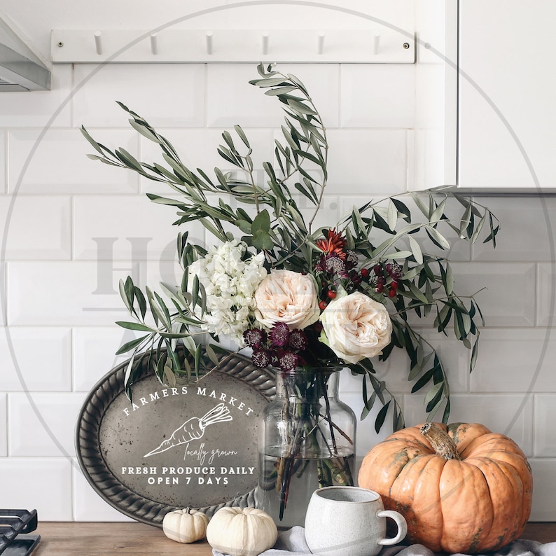 May include: A rustic farmhouse style sign with the text "Farmers Market Locally Grown Fresh Produce Daily Open 7 Days" and a hand-drawn carrot illustration. The sign is displayed on a wooden table with a vase of flowers, a pumpkin, and two small pumpkins.