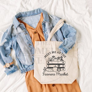 Pu&ograve; includere: Una borsa tote in tela bianca con testo nero che recita "Meet me at the Farmers Market". La borsa presenta un'illustrazione in bianco e nero di una cassa piena di vari frutti e verdure. La borsa &egrave; appoggiata su una superficie bianca con un vestito marrone e una giacca di jeans blu.