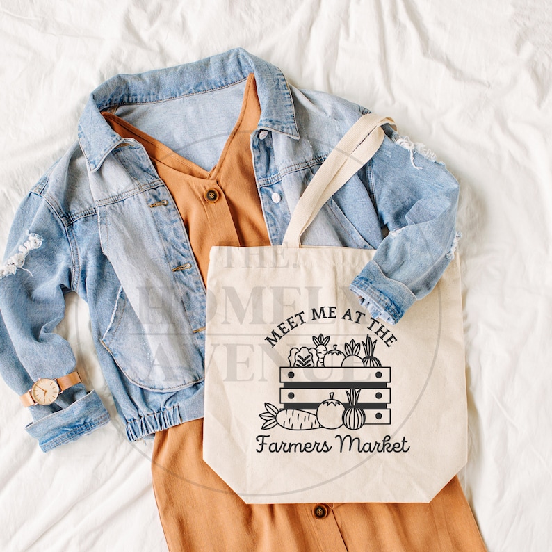Pu&ograve; includere: Una borsa tote in tela bianca con testo nero che recita "Meet me at the Farmers Market". La borsa presenta un'illustrazione in bianco e nero di una cassa piena di vari frutti e verdure. La borsa &egrave; appoggiata su una superficie bianca con un vestito marrone e una giacca di jeans blu.