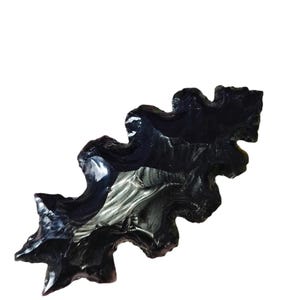 Puede incluir: Un recipiente de vidrio oscuro y translúcido, similar a la obsidiana, con un borde muy irregular, ondulado y dentado. El objeto presenta un centro profundo y hueco con paredes interiores lisas y reflectantes y una superficie exterior.