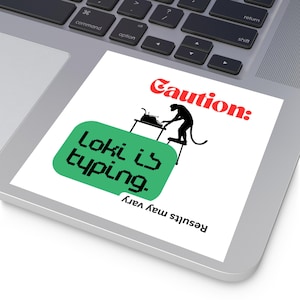 May include: A white sticker on a laptop with the text "Caution: Loki is typing. Results may vary." The sticker features a black silhouette of a monkey typing on a typewriter, with a green speech bubble.