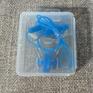 May include: A clear plastic case containing blue swimming accessories. The case holds a pair of earplugs and a nose clip, connected by a blue cord. The items are designed for water activities.