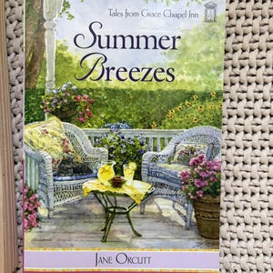 May include: A paperback book titled "Summer Breezes" by Jane Orcutt. The cover art depicts a porch scene with wicker chairs, a small table with lemonade, and potted flowers. The background shows a lush garden.
