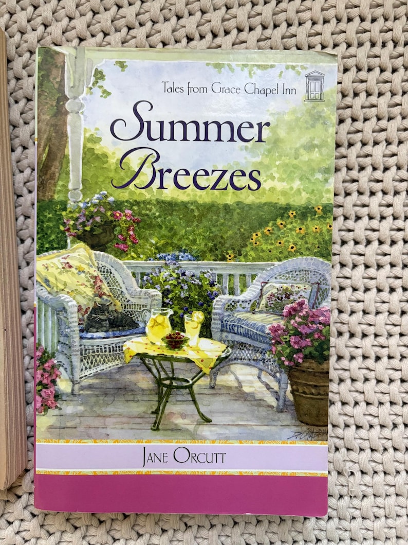 May include: A paperback book titled "Summer Breezes" by Jane Orcutt. The cover art depicts a porch scene with wicker chairs, a small table with lemonade, and potted flowers. The background shows a lush garden.