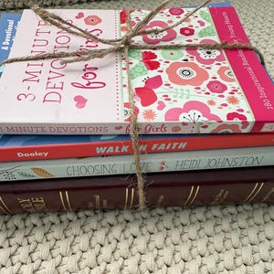 May include: A stack of books tied with twine. The top book is titled "3-Minute Devotions for Girls" with a pink cover and floral design. Below it are books titled "Walk in Faith", "Choosing Love", and a burgundy Bible.