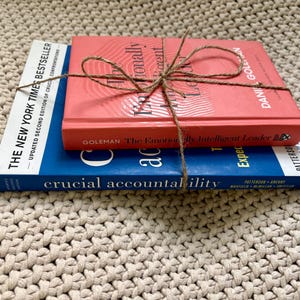 May include: Three books stacked on top of each other, tied with twine. The top book is coral-colored with the title "Emotionally Intelligent Leader." The middle book is blue with the title "Crucial Accountability." The bottom book is white.