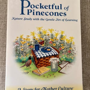 Può includere: Un libro intitolato "Pocketful of Pinecones" di Karen Andreola. La copertina presenta una scena di picnic con un cestino, un uccello e un barattolo. Il sottotitolo del libro è "Nature Study with the Gentle Art of Learning". La parte inferiore del libro recita "A Story for Mother Culture."