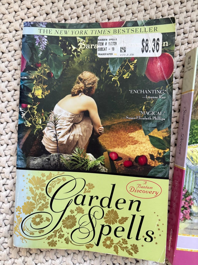 May include: A paperback book titled "Garden Spells" by Sarah Addison Allen. The cover features a woman in a garden setting with red apples. The book is a New York Times bestseller and includes quotes from Luanne Rice and Susan Elizabeth Phillips.
