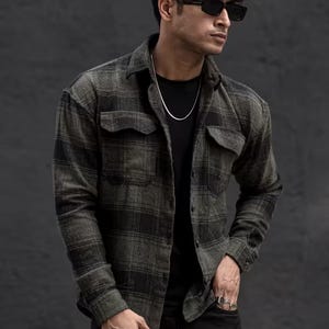 May include: A green and black plaid flannel shirt with two chest pockets. The long-sleeved shirt is buttoned and worn open over a black t-shirt. The person is wearing black pants and sunglasses.