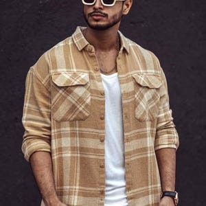 May include: A tan plaid button-down shirt with two chest pockets. The shirt is worn over a white t-shirt and paired with cream-colored pants. The shirt has a classic collar and buttoned cuffs.