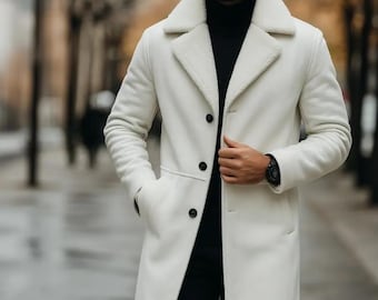 May include: A long, off-white coat with a shearling collar and black buttons. The coat is worn over a black turtleneck and black pants. A black watch is visible on the wrist. The coat has a classic, tailored design.