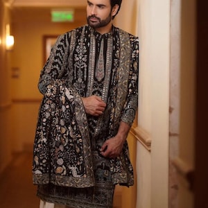 Man wearing an opulent black Mughal-style kurta with intricate multicolor embroidery, paired with cream Patiala trousers and a heavily embellished dupatta featuring animal and floral motifs—ideal for grooms, traditional South Asian weddings, and cultural festivities.