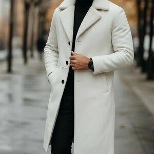 May include: A long, off-white coat with a shearling collar and lining. The coat has a classic design with a button closure and pockets. The person is wearing a black turtleneck and black pants. A black watch is visible on the wrist.