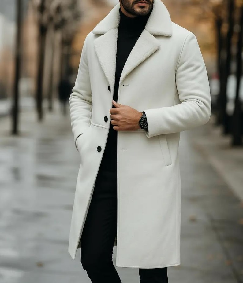 May include: A long, off-white coat with a shearling collar and lining. The coat has a classic design with a button closure and pockets. The person is wearing a black turtleneck and black pants. A black watch is visible on the wrist.