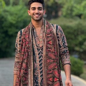 May include: A person wearing a dark green and red paisley patterned outfit, including a long coat and scarf. The outfit has intricate floral and paisley designs in shades of red, brown, and gold.