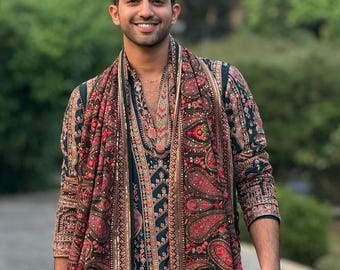 Pode incluir: Uma pessoa vestindo um traje com estampa paisley verde escuro e vermelho, incluindo um casaco longo e um lenço. O traje tem intrincados desenhos florais e paisley em tons de vermelho, marrom e dourado.