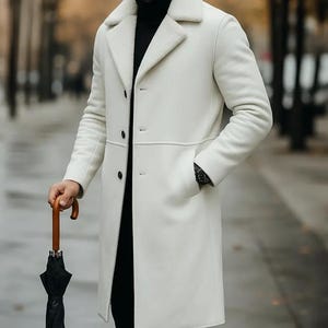 May include: A white, long coat with a shearling collar and lining. The coat has a three-button closure and two pockets. The person is holding a black umbrella with a wooden handle. The person is wearing a black turtleneck and black pants.