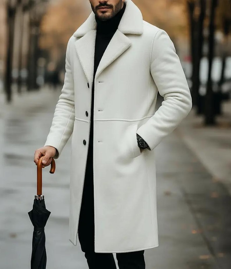 May include: A white, long coat with a shearling collar and lining. The coat has a three-button closure and two pockets. The person is holding a black umbrella with a wooden handle. The person is wearing a black turtleneck and black pants.