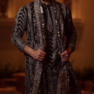 Man wearing an opulent black Mughal-style kurta with intricate multicolor embroidery, paired with cream Patiala trousers and a heavily embellished dupatta featuring animal and floral motifs—ideal for grooms, traditional South Asian weddings, and cultural festivities.