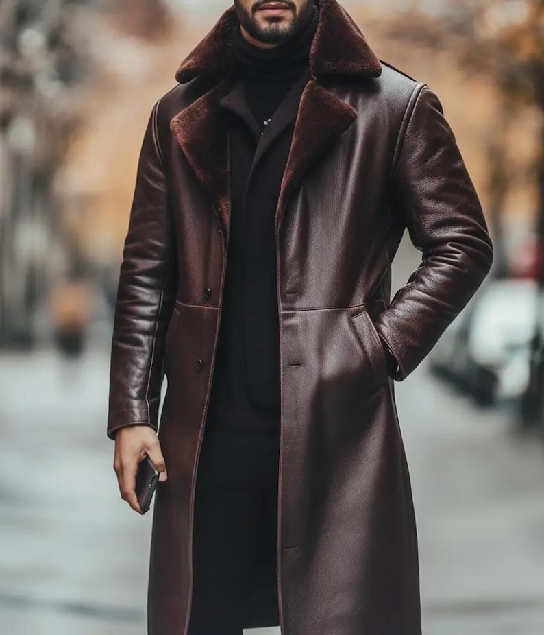 May include: A long, dark brown leather coat with a faux fur collar. The coat has a button-down front, pockets, and a tailored fit. The coat is worn over a black turtleneck and black pants. The person is holding a black phone.