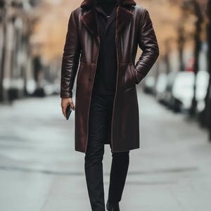 May include: A long, burgundy leather coat with a faux fur collar. The coat is buttoned and worn over a black turtleneck and black pants. The person is holding a phone and wearing black shoes. The background is a blurred city street.