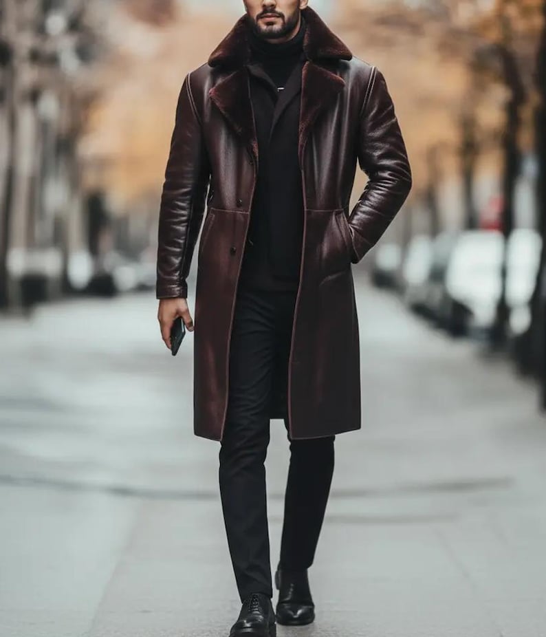 May include: A long, burgundy leather coat with a faux fur collar. The coat is buttoned and worn over a black turtleneck and black pants. The person is holding a phone and wearing black shoes. The background is a blurred city street.