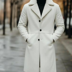 May include: A long, white coat with a shearling collar and black buttons. The coat has two front pockets and a tailored fit. The person in the image is wearing a black turtleneck underneath the coat.