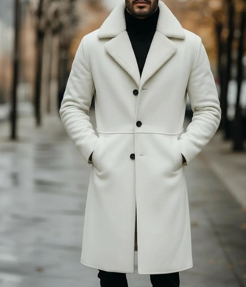 May include: A long, white coat with a shearling collar and black buttons. The coat has two front pockets and a tailored fit. The person in the image is wearing a black turtleneck underneath the coat.
