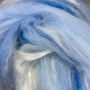 Puede incluir: Primer plano de un haz de fibra azul claro y blanco con hebras de purpurina plateada. La textura suave y esponjosa de la fibra es visible, con diferentes tonos de azul y blanco mezcl&aacute;ndose. Este material se utiliza probablemente para proyectos de artesan&iacute;a o textiles.