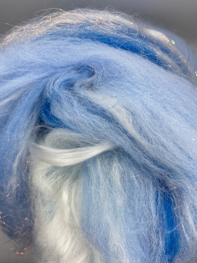 Puede incluir: Primer plano de un haz de fibra azul claro y blanco con hebras de purpurina plateada. La textura suave y esponjosa de la fibra es visible, con diferentes tonos de azul y blanco mezcl&aacute;ndose. Este material se utiliza probablemente para proyectos de artesan&iacute;a o textiles.