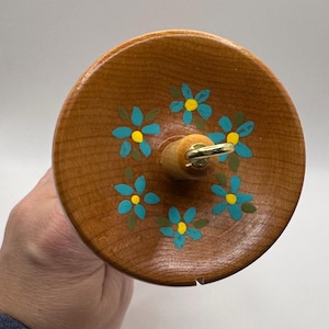 May include: A wooden drop spindle with a round top featuring hand-painted blue and yellow flowers. The spindle has a wooden shaft and a gold-colored metal ring. The item is designed for spinning yarn.