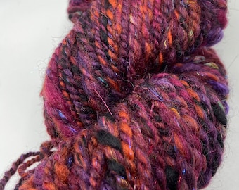 May include: A close-up of a skein of yarn in shades of purple, orange, and black. The yarn has a thick, textured appearance with strands of shimmering, iridescent fibers throughout. This yarn is suitable for knitting and crochet projects.
