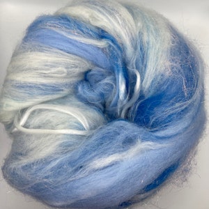 Puede incluir: Una madeja de fibra azul y blanca con hebras plateadas. La fibra es suave y esponjosa, con tonos azul claro y oscuro. La textura es delicada y aireada, adecuada para manualidades. La madeja est&aacute; bien enrollada.