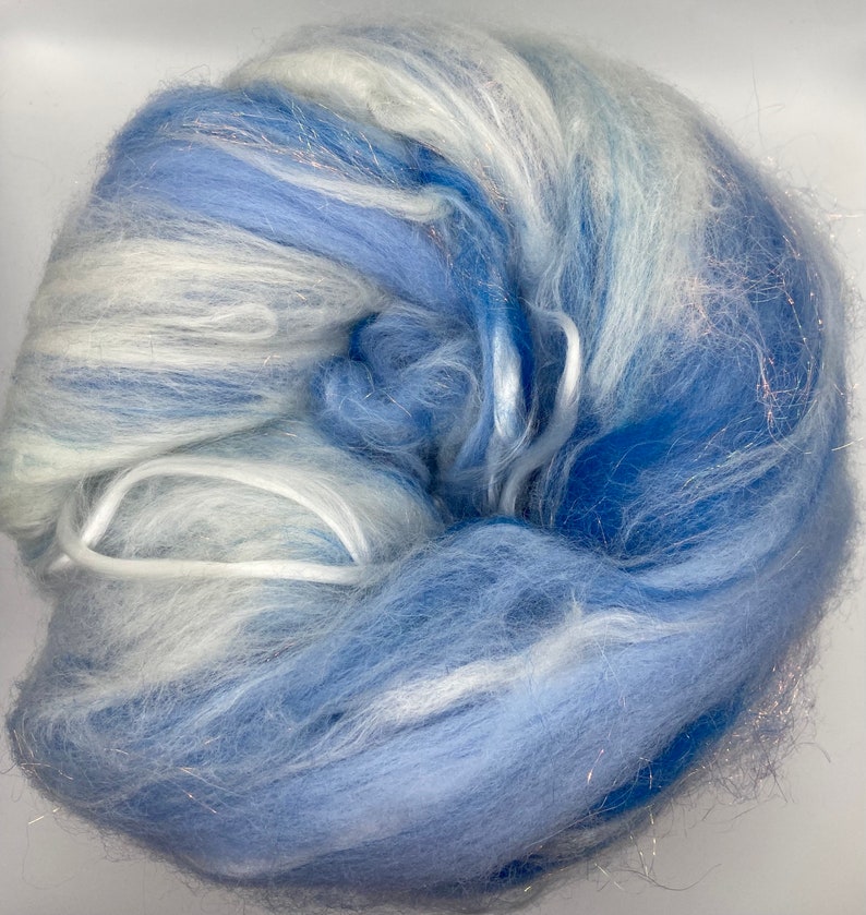 Puede incluir: Una madeja de fibra azul y blanca con hebras plateadas. La fibra es suave y esponjosa, con tonos azul claro y oscuro. La textura es delicada y aireada, adecuada para manualidades. La madeja est&aacute; bien enrollada.