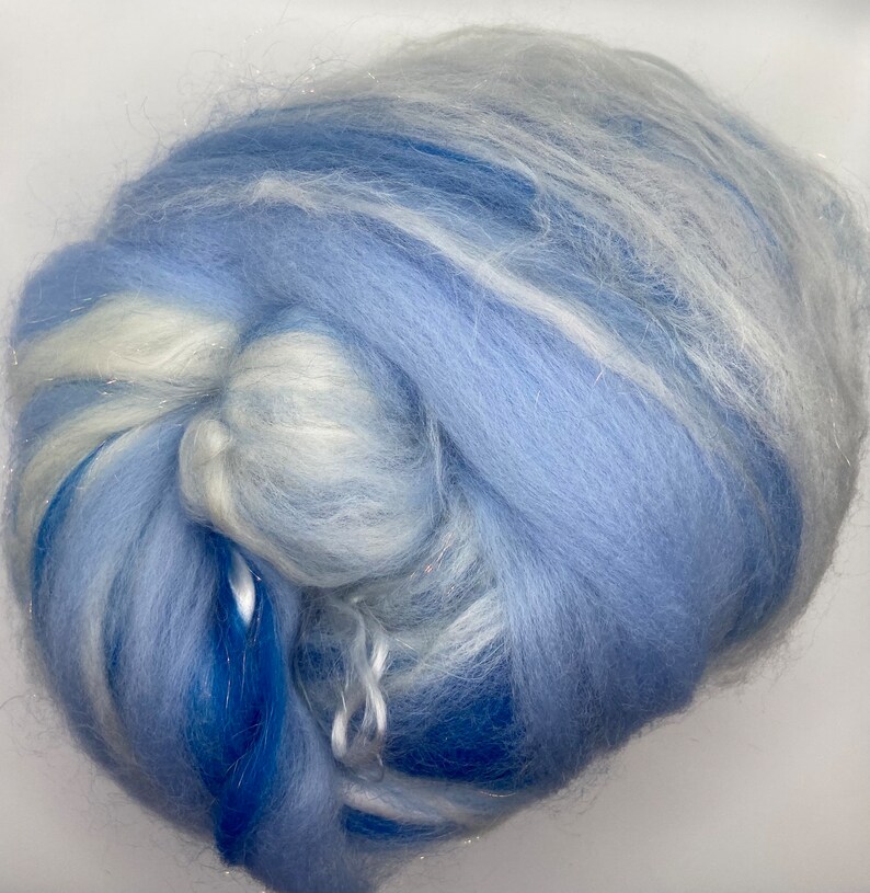 Puede incluir: Primer plano de una nube de fibra en tonos azules y blancos. La fibra es suave y esponjosa, con hebras de purpurina plateada. Los colores se mezclan, creando un efecto so&ntilde;ador y et&eacute;reo. Esta fibra se utiliza probablemente para manualidades.
