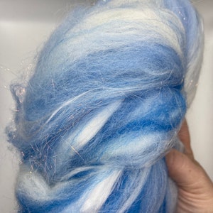 Puede incluir: Un paquete de fibra azul y blanca con una textura suave y esponjosa. La fibra tiene un efecto degradado, que va de un azul intenso a un tono m&aacute;s claro, con vetas blancas. Se intercalan finos hilos brillantes.