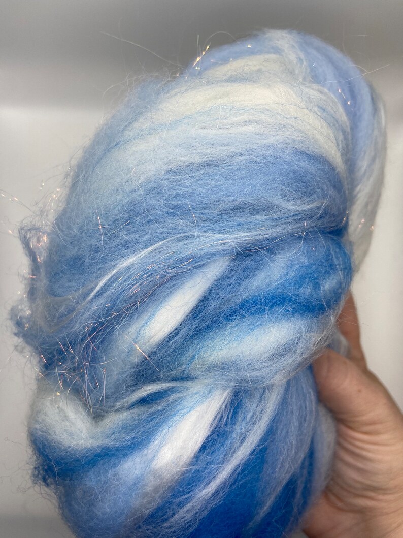 Puede incluir: Un paquete de fibra azul y blanca con una textura suave y esponjosa. La fibra tiene un efecto degradado, que va de un azul intenso a un tono m&aacute;s claro, con vetas blancas. Se intercalan finos hilos brillantes.