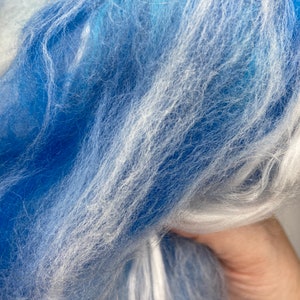 Puede incluir: Primer plano de un manojo de fibra azul y blanca. Las fibras son suaves y esponjosas, con una mezcla de tonos azules claros y oscuros, y reflejos blancos. La textura parece delicada y aireada, lo que sugiere un material artesanal como hilo o roving.
