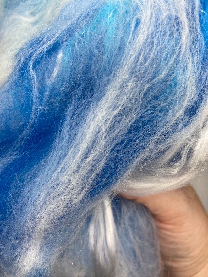 Puede incluir: Primer plano de un manojo de fibra azul y blanca. Las fibras son suaves y esponjosas, con una mezcla de tonos azules claros y oscuros, y reflejos blancos. La textura parece delicada y aireada, lo que sugiere un material artesanal como hilo o roving.