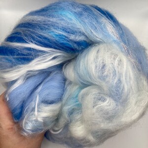 Puede incluir: Una masa de fibra azul y blanca. La fibra es una mezcla de azules claros y oscuros, con vetas blancas y un toque de plata. La textura parece suave y esponjosa, adecuada para hilar u otras artes de la fibra.