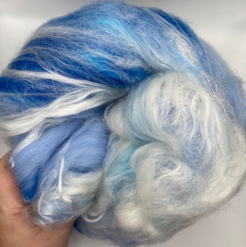 Puede incluir: Una masa de fibra azul y blanca. La fibra es una mezcla de azules claros y oscuros, con vetas blancas y un toque de plata. La textura parece suave y esponjosa, adecuada para hilar u otras artes de la fibra.