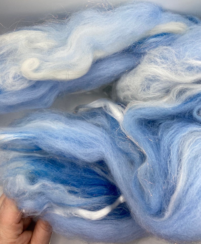 Puede incluir: Primer plano de un mont&oacute;n de fibra suave y esponjosa en tonos azules y blancos. La fibra tiene una textura delicada y aireada, con hebras de purpurina plateada entretejidas. El efecto general es et&eacute;reo y similar a una nube, perfecto para manualidades.