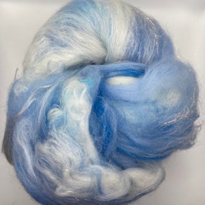 Puede incluir: Un ovillo de fibra en tonos azules y blancos, con un brillo sutil. La fibra parece suave y esponjosa, lo que sugiere que podr&iacute;a usarse para manualidades o proyectos textiles. La apariencia general es delicada y aireada.