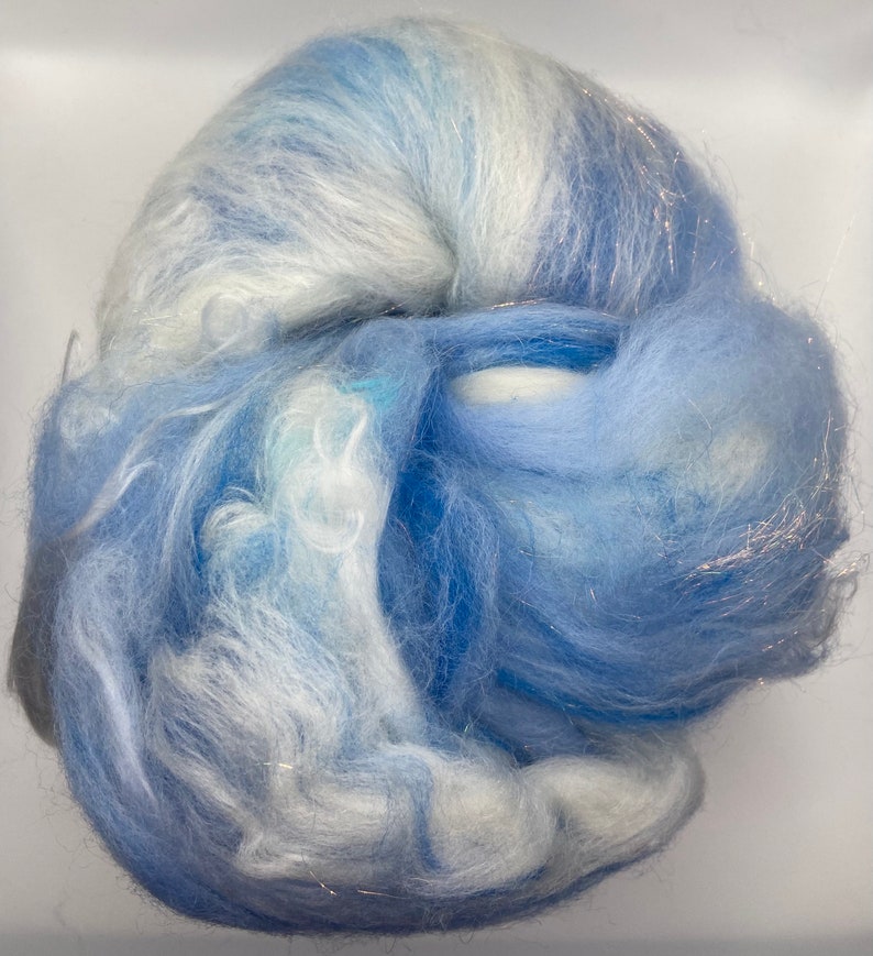 Puede incluir: Un ovillo de fibra en tonos azules y blancos, con un brillo sutil. La fibra parece suave y esponjosa, lo que sugiere que podr&iacute;a usarse para manualidades o proyectos textiles. La apariencia general es delicada y aireada.