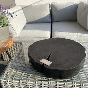 May include: A black wooden serving tray with a natural wood grain pattern and a silver metal accent. The tray is round and has a slightly uneven edge. It is sitting on a white wicker table with a glass top.