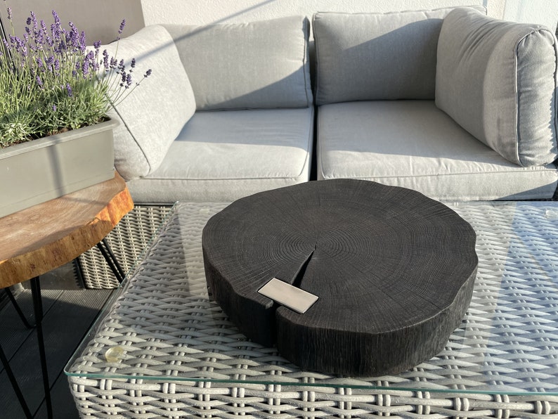 May include: A black wooden serving tray with a natural wood grain pattern and a silver metal accent. The tray is round and has a slightly uneven edge. It is sitting on a white wicker table with a glass top.