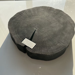 May include: A large, round, black wooden serving board with a natural wood grain pattern. The board has a small, rectangular, silver metal insert in the center.